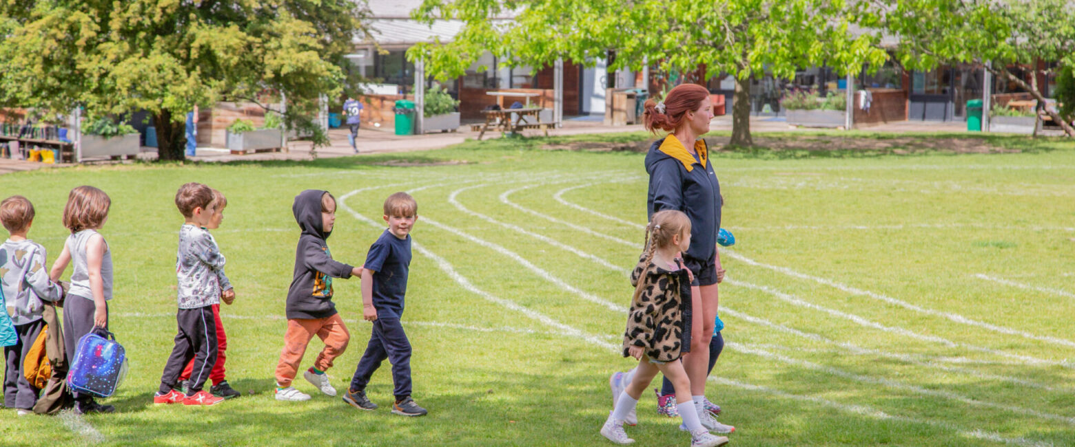 Lower School students from The King Alfred School in Hampstead walk across a field marked with athletics tracks alongside a teacher