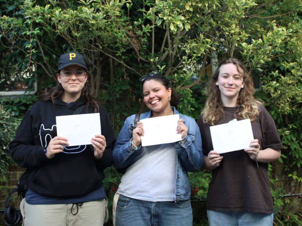 Three students from The King Alfred School smile and hold their A level exam results certificates in front of a bush