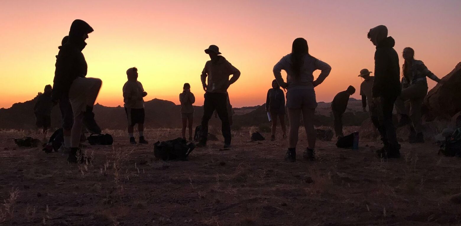 Students from The King Alfred School in silhouette standing in the Namibian desert as the sun sets