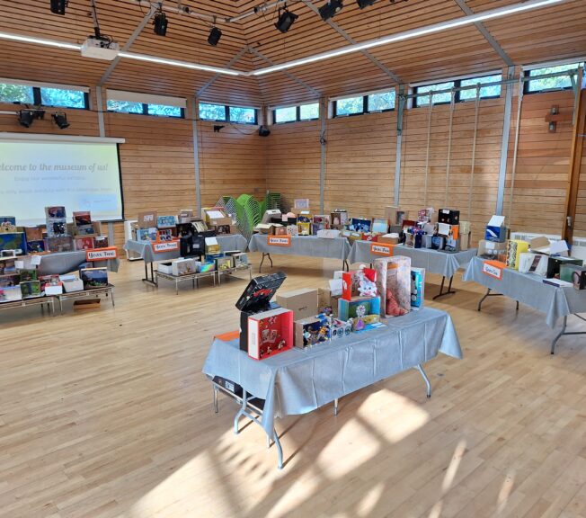 Lines of tables topped with decorated shoeboxes inside a wooden school gym, with a sign that reads 'welcome to the Museum of Us'.
