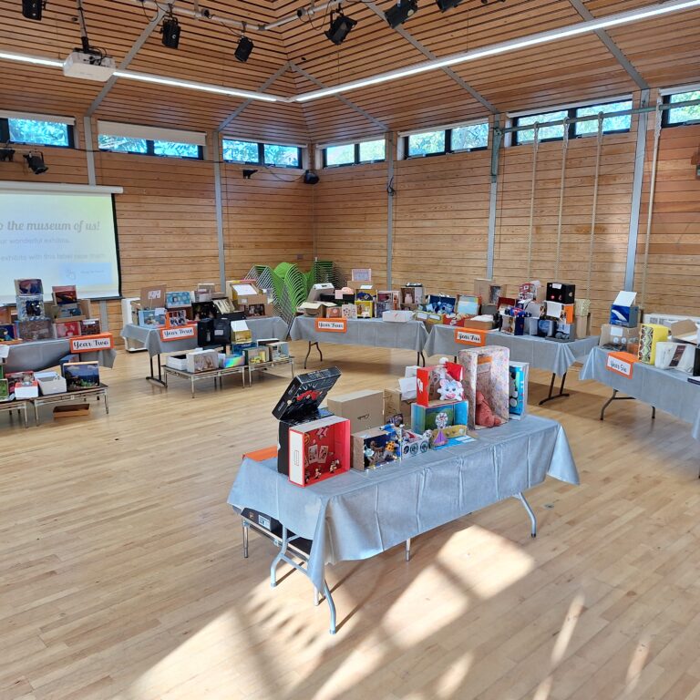 Lines of tables topped with decorated shoeboxes inside a wooden school gym, with a sign that reads 'welcome to the Museum of Us'.