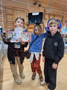 Three young children stand in a line. One holds a decorated shoebox with a cassette player inside connected to pairs of headphones, one of which is worn by another child. The third child holds a decorated shoebox above their head.