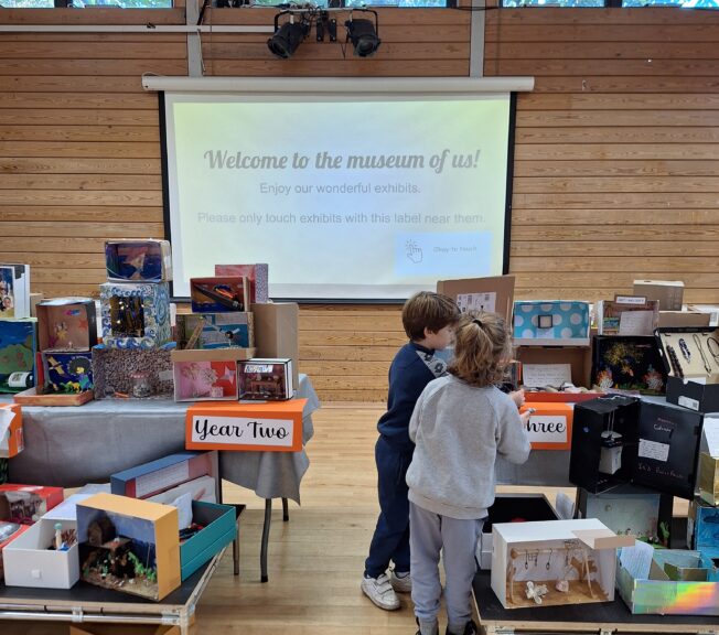 Two young children look at a collection of decorated shoeboxes on a table.