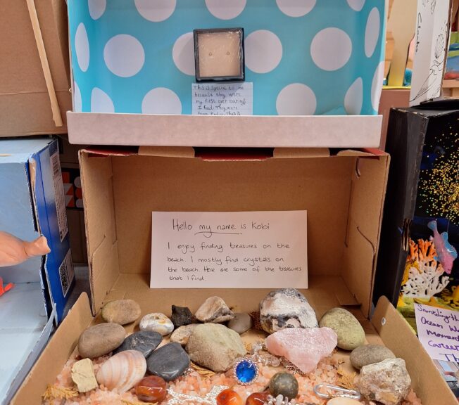 Two shoeboxes on top of one another. One has blue and white spotted paper and a pair of small earrings inside. A note reads: 'This is special to me because they were my first ever earrings that I had. They were from India.' The other box has shells and trinkets inside, with a note that reads: 'Hello my name is Kobi. I enjoy finding treasures on the beach. I mostly find crystals on the beach. Here are some of the treasures that I find.'
