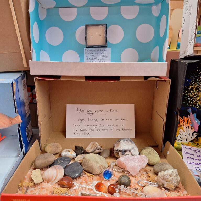 Two shoeboxes on top of one another. One has blue and white spotted paper and a pair of small earrings inside. A note reads: 'This is special to me because they were my first ever earrings that I had. They were from India.' The other box has shells and trinkets inside, with a note that reads: 'Hello my name is Kobi. I enjoy finding treasures on the beach. I mostly find crystals on the beach. Here are some of the treasures that I find.'