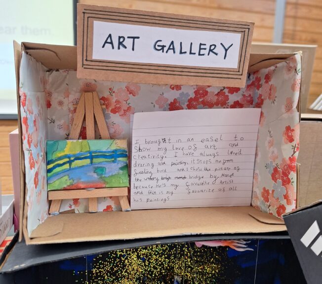 A shoebox covered in floral paper, with a small art easel and paining of a watercolour scene inside. A note reads: 'Art Gallery. I brought in an easel to show my love of art and creativity. I have always loved drawing and painting, it stops me from feeling board and I chose this picture as the watery bridge by Monet because he's my favourite artist and this is my favourite of all his paintings.'