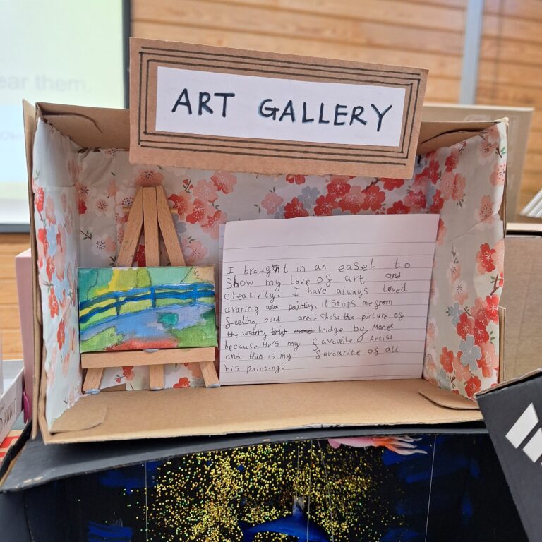 A shoebox covered in floral paper, with a small art easel and paining of a watercolour scene inside. A note reads: 'Art Gallery. I brought in an easel to show my love of art and creativity. I have always loved drawing and painting, it stops me from feeling board and I chose this picture as the watery bridge by Monet because he's my favourite artist and this is my favourite of all his paintings.'