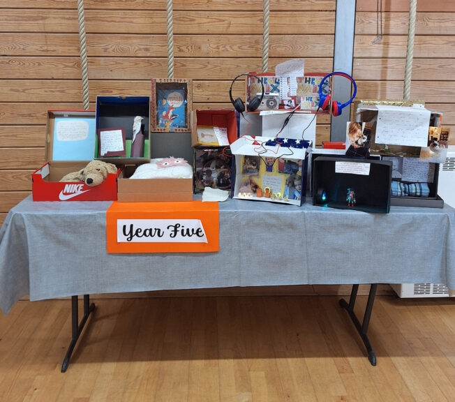 A table of decorated shoeboxes on a table in a wooden school gym. A sign reads 'Year Five'.