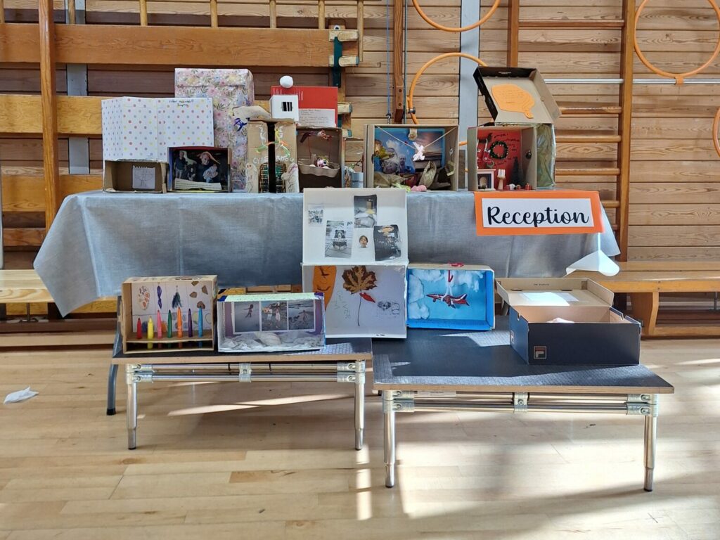 A table of decorated shoeboxes on a table in a wooden school gym. A sign reads 'reception'.