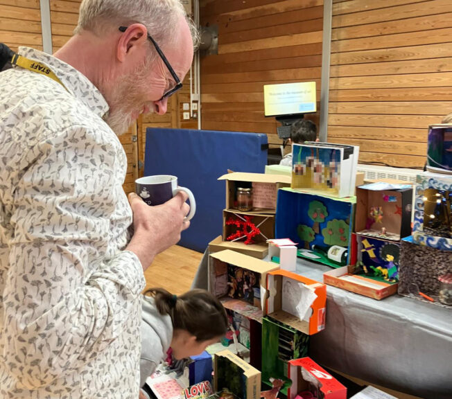 A man smiles as he takes in a display of decorated shoeboxes, with a young girl leaning in for a closer look