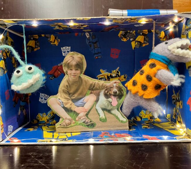 The inside of a shoebox decorated with Transformers paper, a toy dinosaur and alien, and a picture of a smiling boy with a happy dog.