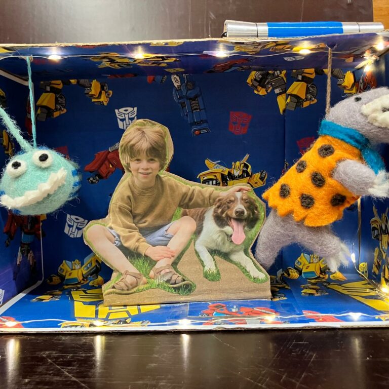 The inside of a shoebox decorated with Transformers paper, a toy dinosaur and alien, and a picture of a smiling boy with a happy dog.