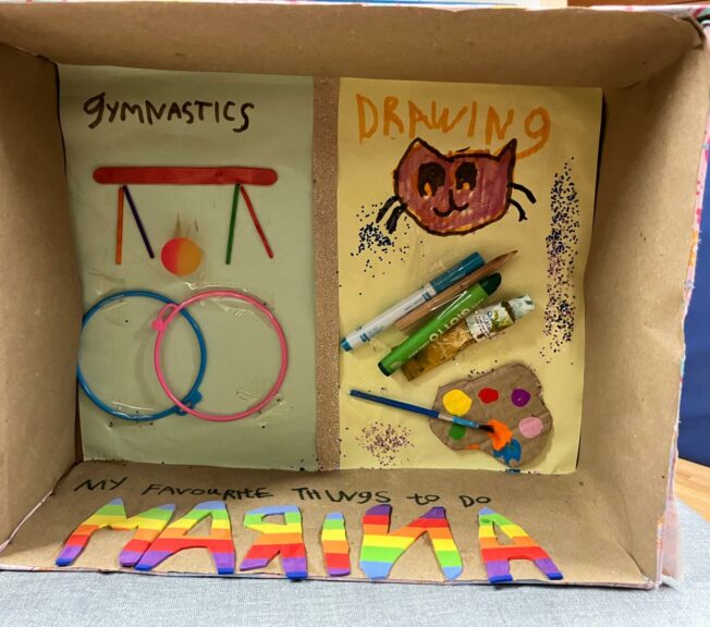 A shoebox on its side with two pictures inside, one depicting gymnastics and one depicting 'drawing', with a paint palette and pens. A note reads: 'My favourite things to do'.