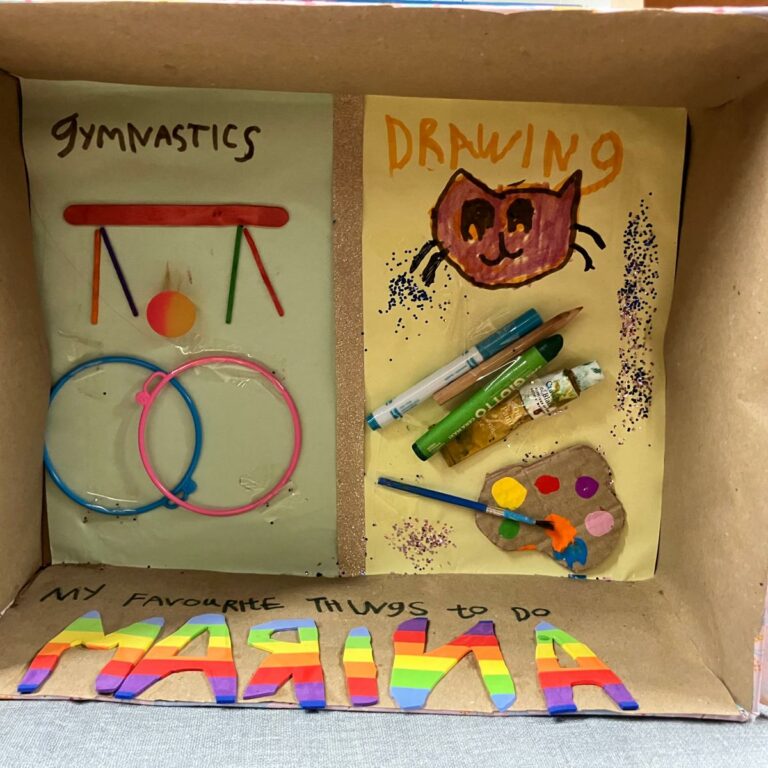 A shoebox on its side with two pictures inside, one depicting gymnastics and one depicting 'drawing', with a paint palette and pens. A note reads: 'My favourite things to do'.