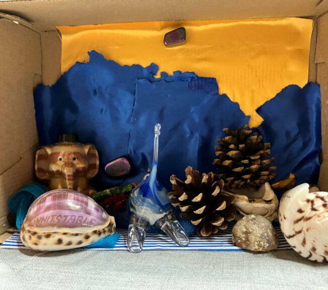 A shoebox on its side, painted blue and orange and decorated with shells and pinecones.