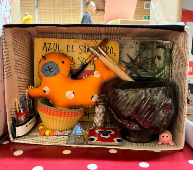 A shoebox on its side, decorated inside with pages from a book, a bank note, paintbrushes and pots.