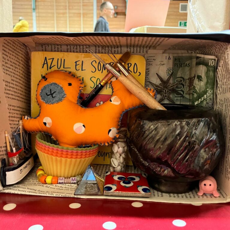 A shoebox on its side, decorated inside with pages from a book, a bank note, paintbrushes and pots.