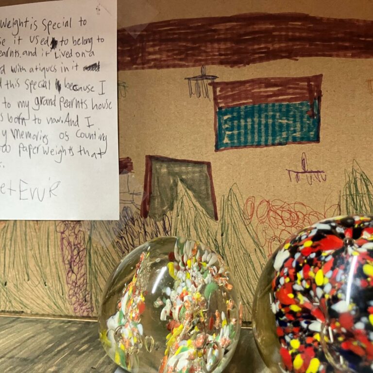 A shoebox decoated with drawings on the inside alongside two paper weights. A note reads: 'This paperweight is special to me because it used to belong to my grandparents and it lived on a glass cupboard with antiques in it. Why I find this special is because I have been to my grandparents' house since I was born to now. And I have lovely memories of counting all 1,000 paperweights that they collect.'