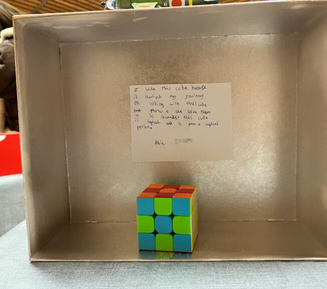 The inside of a shoebox painted silver, with a Rubik's Cube alongside a note that reads: 'I like this cube because it started my journey n cubing with this cube and now I can solved them in 30 seconds. This cube is logical and I am a logical person.'