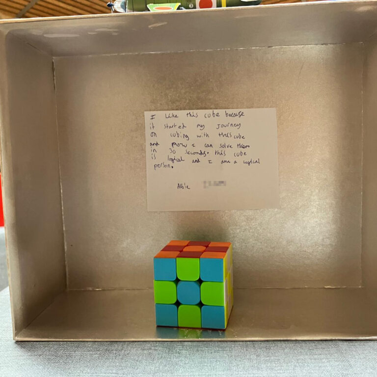 The inside of a shoebox painted silver, with a Rubik's Cube alongside a note that reads: 'I like this cube because it started my journey n cubing with this cube and now I can solved them in 30 seconds. This cube is logical and I am a logical person.'