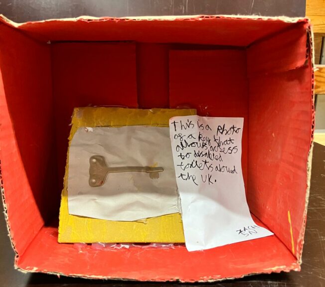 A shoebox on its side painted red inside. There is a photograph of a key and a note that reads: 'This is a photo of a key that allows access to disabled toilets around the UK'