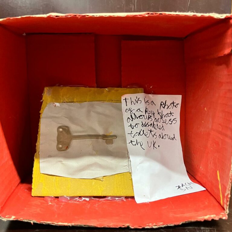 A shoebox on its side painted red inside. There is a photograph of a key and a note that reads: 'This is a photo of a key that allows access to disabled toilets around the UK'
