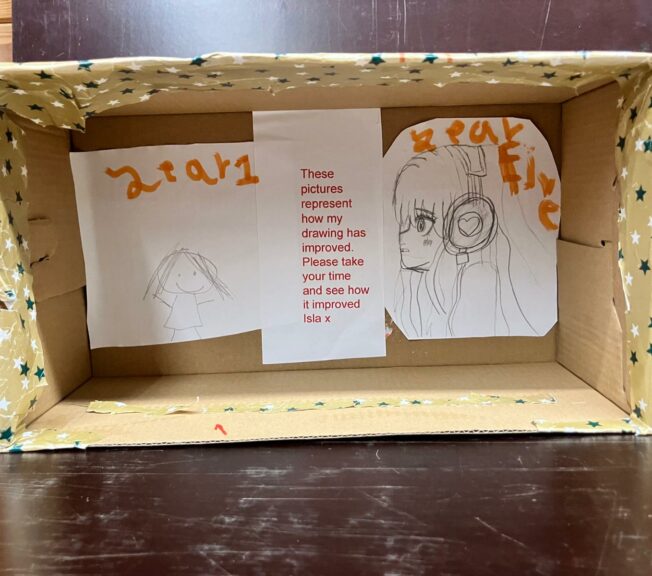 A shoebox on its side covered in spotty wrapping paper. There are two pencil drawings side by side, with a note that reads: 'These pictures represent how my drawing has improved. Please take your time and see how is improved.'