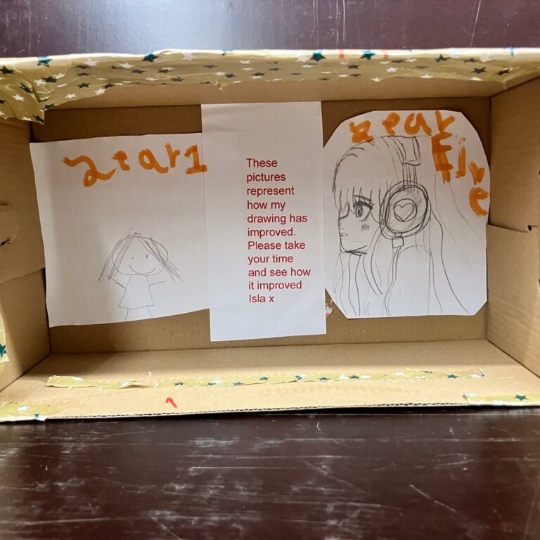 A shoebox on its side covered in spotty wrapping paper. There are two pencil drawings side by side, with a note that reads: 'These pictures represent how my drawing has improved. Please take your time and see how is improved.'
