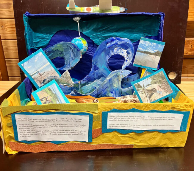 A shoebox painted blue and yellow to look like the beach and sea with waves. There are also shells and pictures of children on a beach, with a note that reads: 'This year my family and I went travelling around the world for four moths. We went to Costa Rica, Panama, USA, California, Fiji, New Zealand, Australia and Hong Kong. During our travels my brother and I loved finding shells like these on the tropical beaches.'