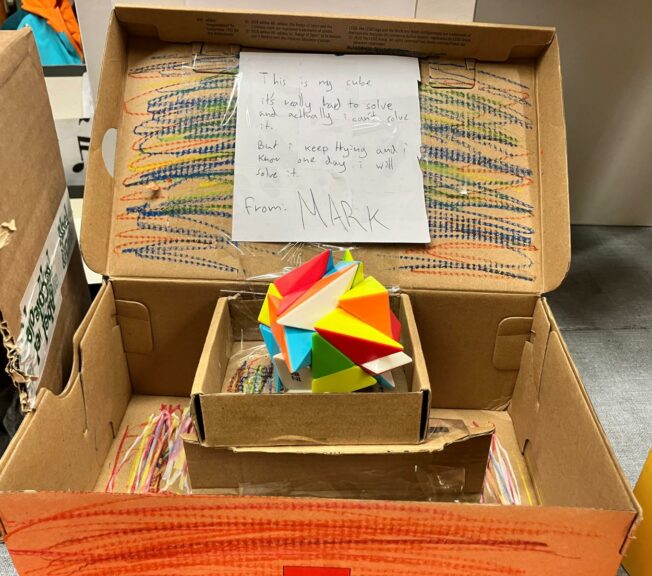 A decorated shoebox with a puzzle block inside. A note reads: 'This is my cube, it's really hard to solve and actually I can't solve it. But I keep trying and I know one day I will solve it.'