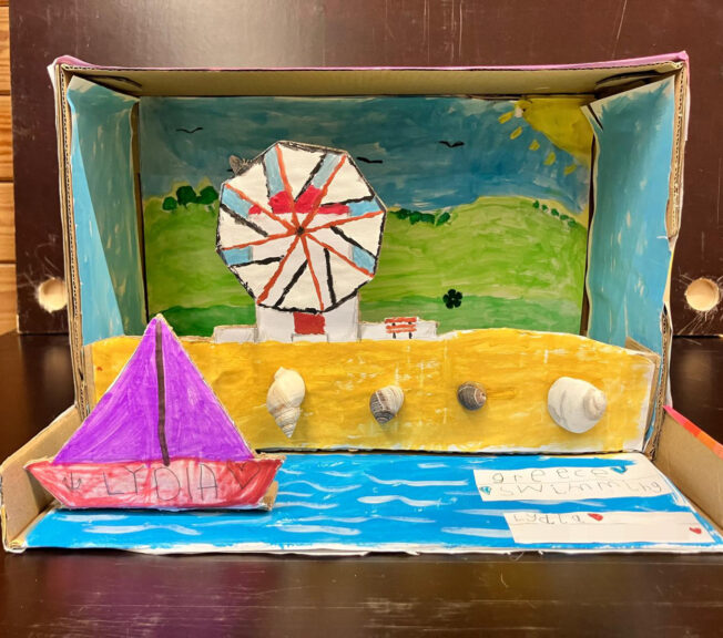 The inside of a shoebox decorated to look like a coastal scene, with a boat in the water, shells on the beach and a windmill in the background.