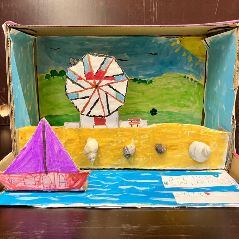 The inside of a shoebox decorated to look like a coastal scene, with a boat in the water, shells on the beach and a windmill in the background.