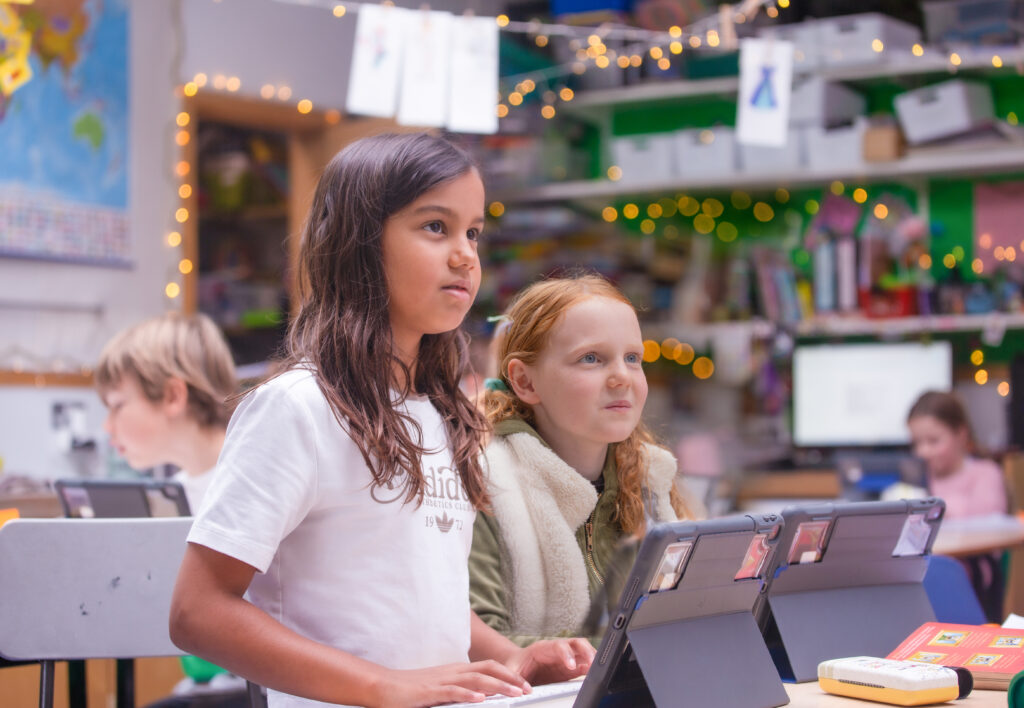 Two female students sit at a desk with iPads on the desk in a school classroom, with fairy lights in the background