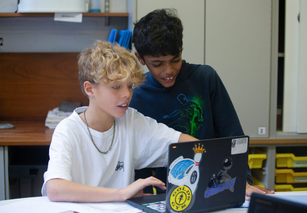 Two male students, one white and one Asian, look at a laptop in a school classroom