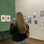 A female Sixth Form student from The King Alfred School pictured from behind taking a photo of a photography exhibition display on her smartphone