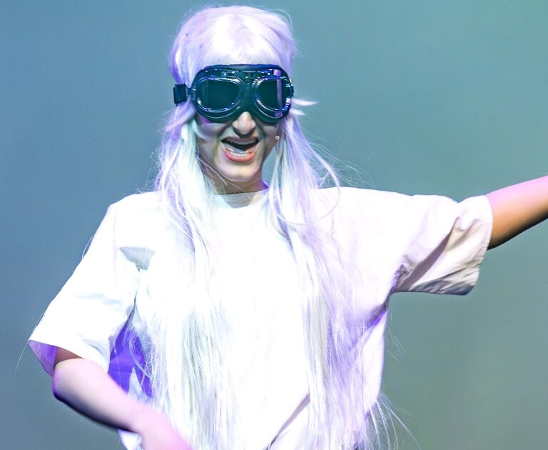 A performer dressed as a scientist in a white lab coat and goggles, holding a skull