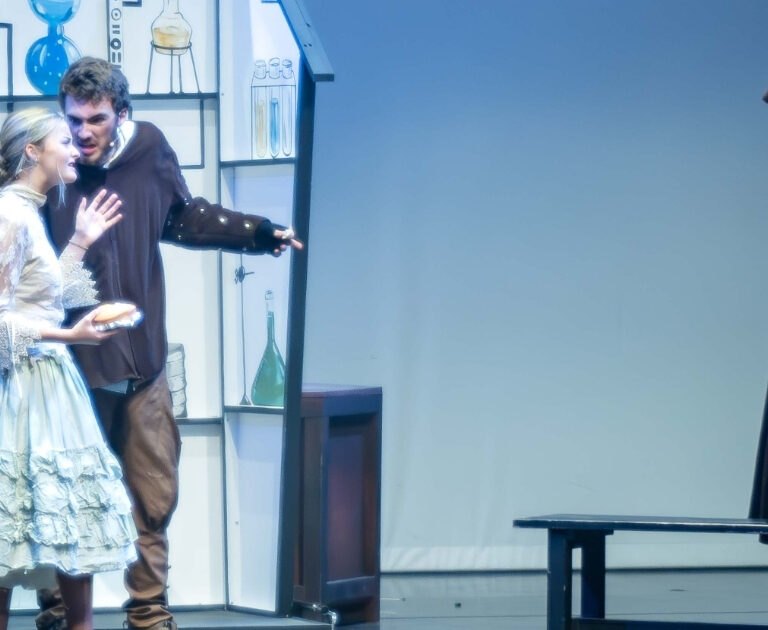 Actors on stage. One is dressed in a black hooded cape, whilst the others look scared