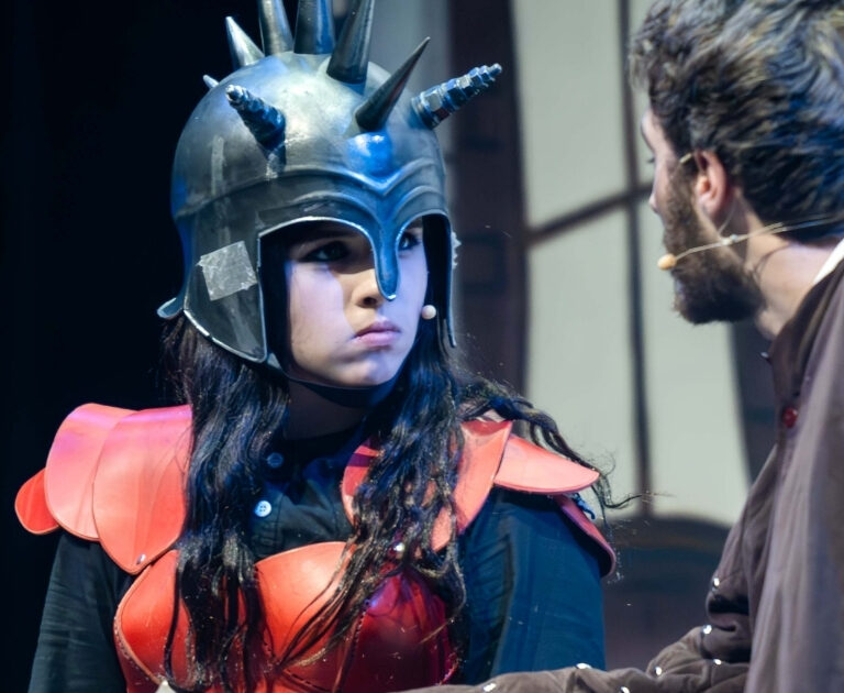 A female actor, wearing a spiky helmet, glowers at another