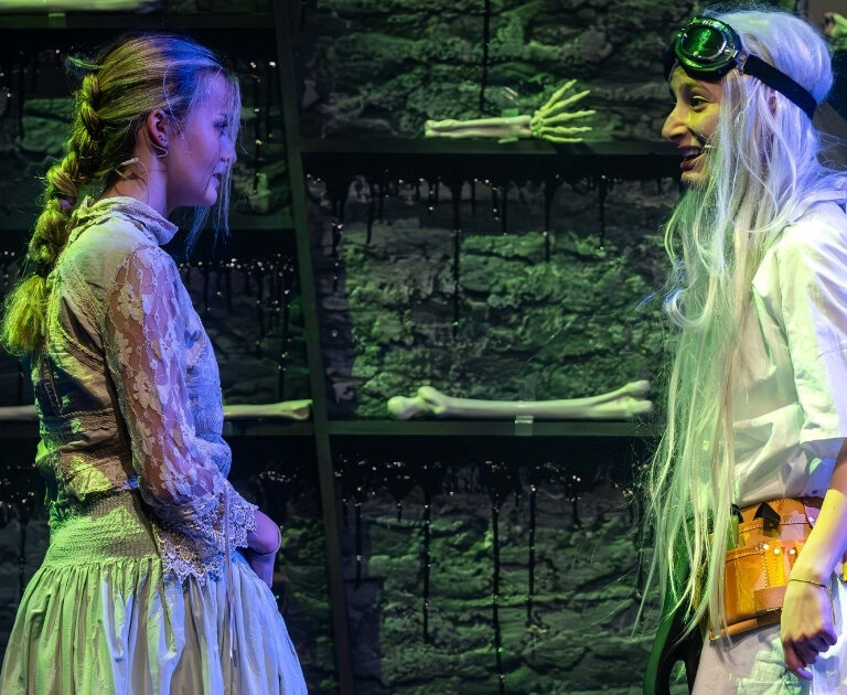 Two actors face each other one stage, with shelves of bones in the backgroun