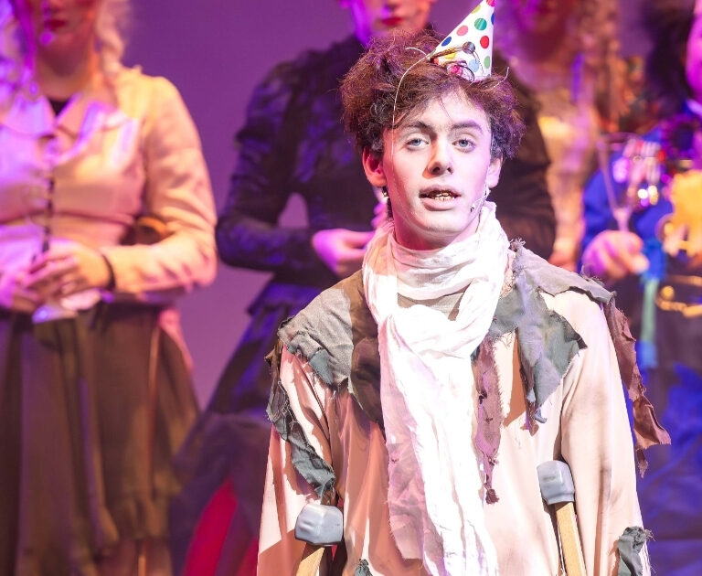 An actor wears a party hat and walks with crutches on stage