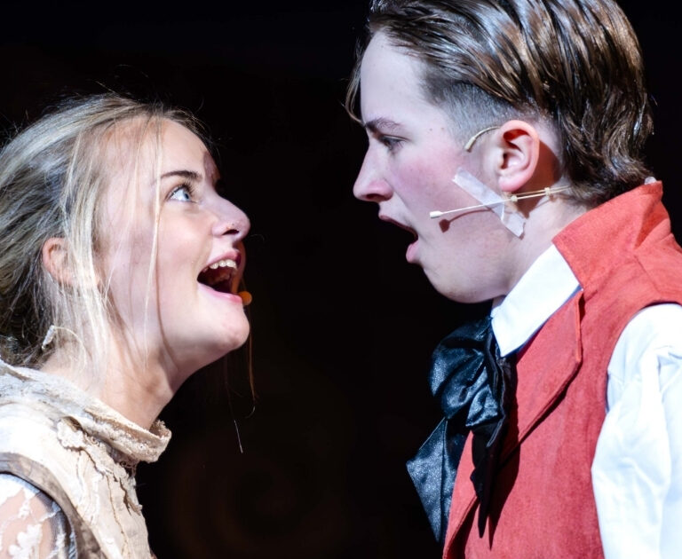 Two actors face one another whilst singing, their faces full of emotion