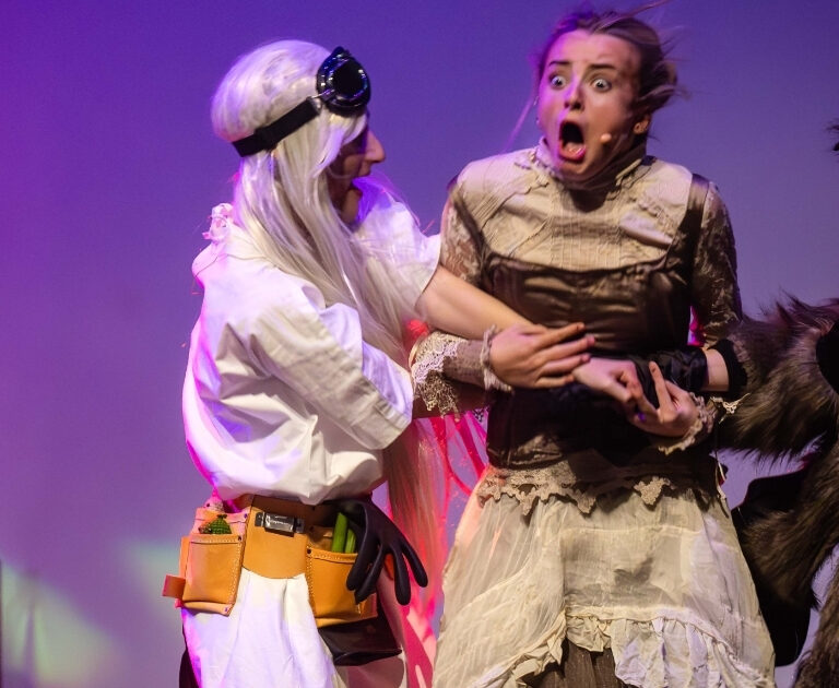 A blonde actor looks surprised as she is seized by fellow performers