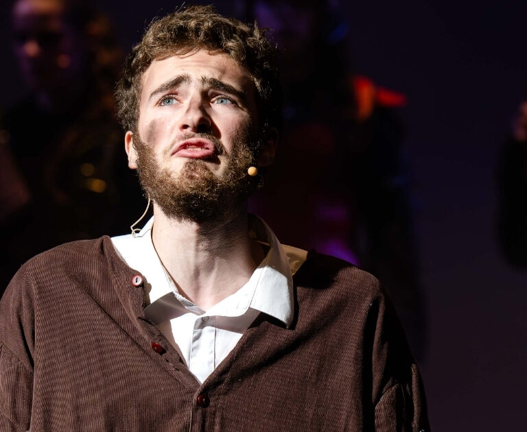 A beadered male actor in a brown smock over a white shirt, with dirt on his face