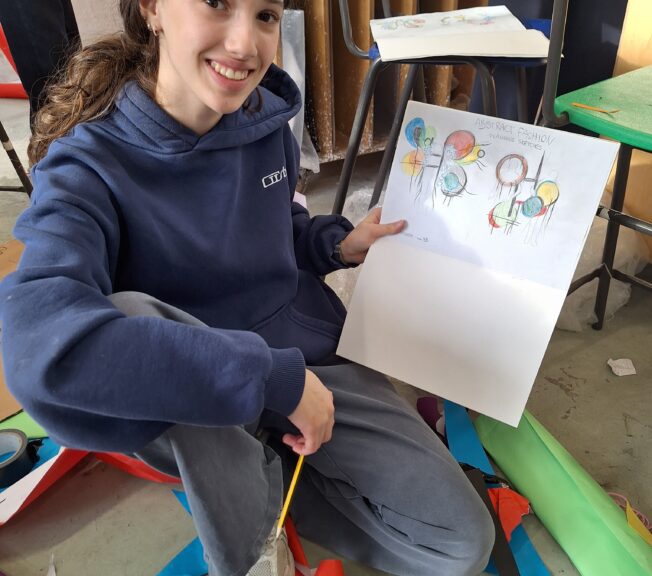 A student smiles as she holds up a sketch of her abstract fashion design