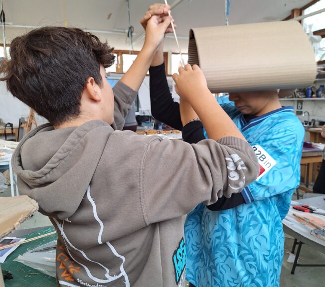 Two male students create a wearable sculpture. One has a cylinder of card around his head and the other attaches a stick.