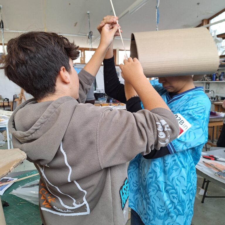 Two male students create a wearable sculpture. One has a cylinder of card around his head and the other attaches a stick.