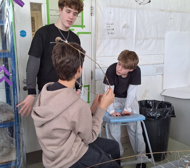 Three students consider their abstract fashion design, with one holding a circle made of sticks.