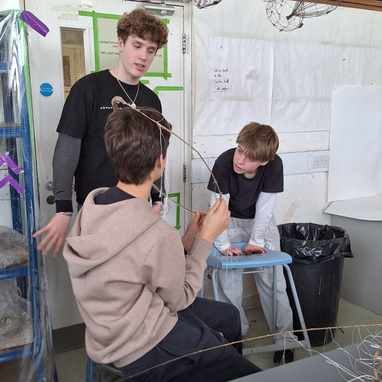 Three students consider their abstract fashion design, with one holding a circle made of sticks.