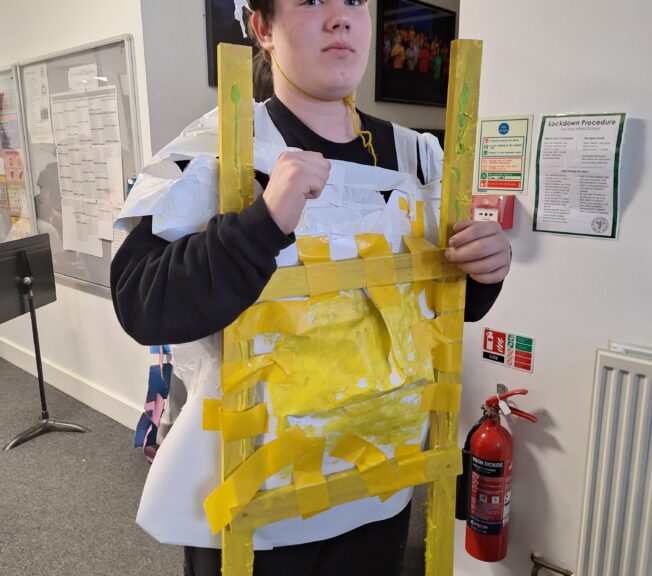A male student wears a white tabard with strips of yellow paper across the front, giving the illusion of a fried egg