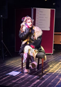 A student performs on stage at The King Alfred School. They are using a mobile phone and wearing a wig and large coat